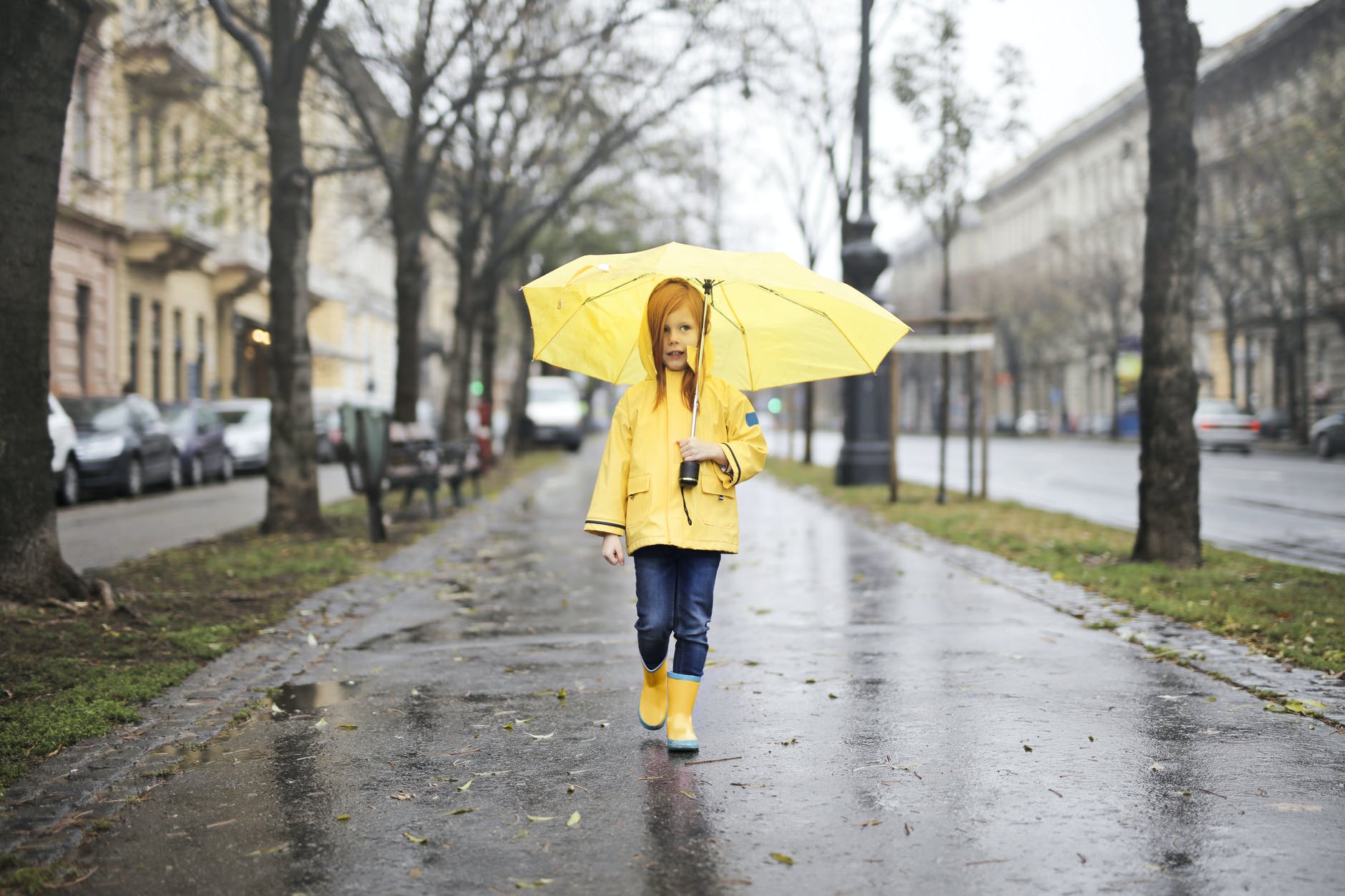 girl in yellow raincoat and yellow boots holding yellow umbrella walking on gray concrete side walk